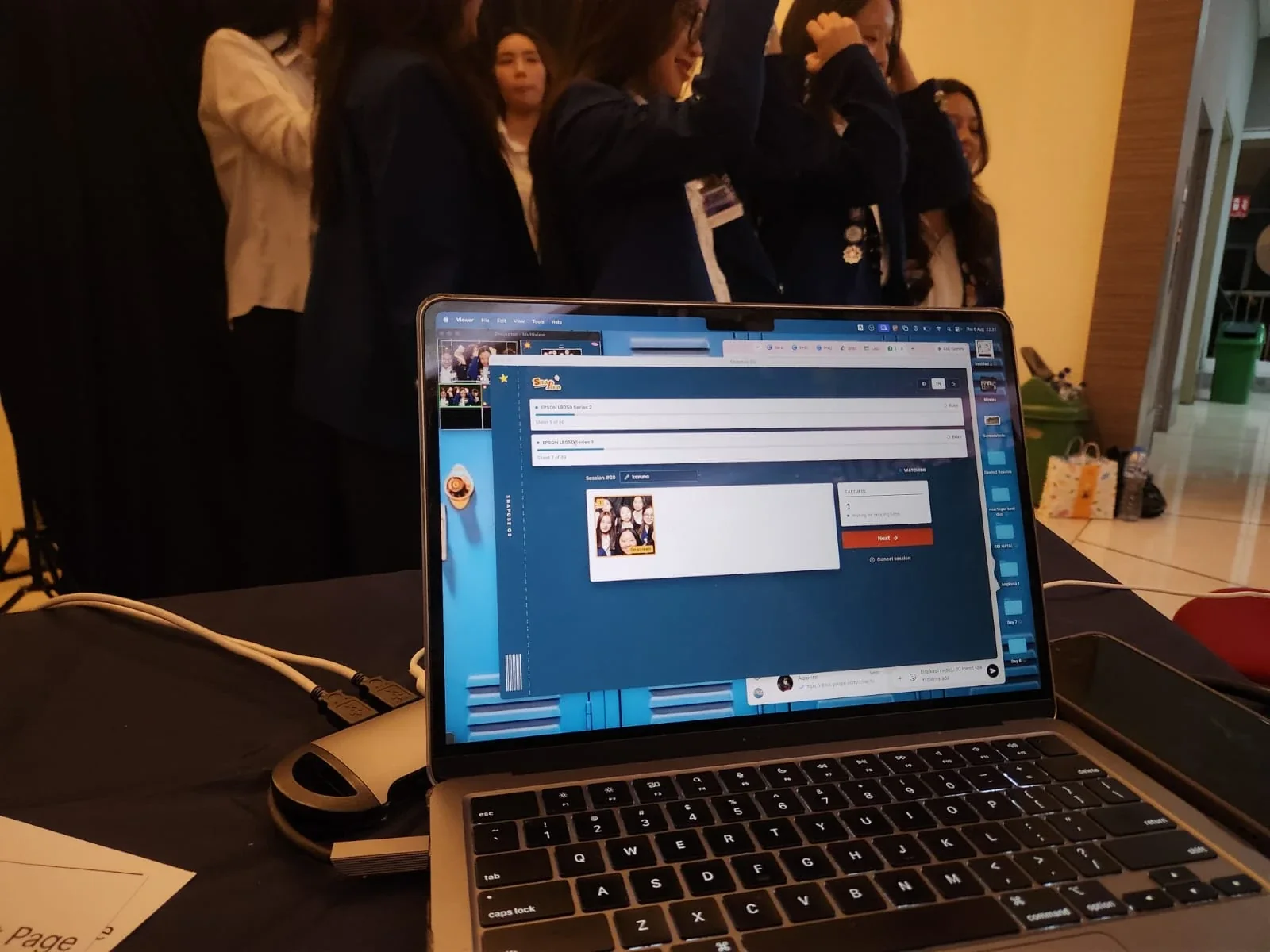 A laptop running Snapose at a live event, two printers in the queue, guests posing behind it